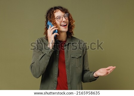 Happy calm fancy young brunet curly man 20s wears khaki shirt hold in hand use talk speak on mobile cell phone conducting pleasant conversation isolated on plain olive green background studio portrait