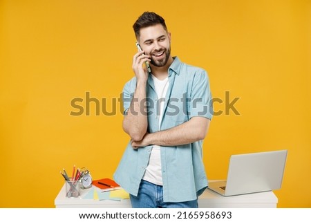 Young successful employee business man in shirt stand work at white office desk with pc laptop talk on mobile cell phone conducting pleasant conversation isolated on yellow background studio portrait