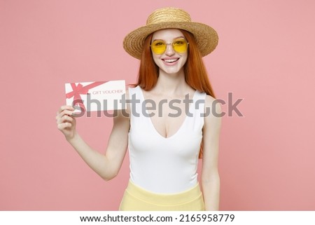 Young smiling nice redhead woman 20s ginger long hair wearing straw hat glasses summer clothes holding gift voucher flyer mock up look camera isolated on pastel pink color background studio portrait