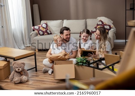 A family unpacks cardboard boxes together after moving into a new apartment. A couple and their children look at old photos in frames, find beautiful keepsakes, tidy up their home.