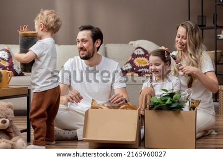 Parents with two small children move into a new apartment, unpack cardboard boxes, take out things, sweet son carries a vase, mom braids her daughter's braids.