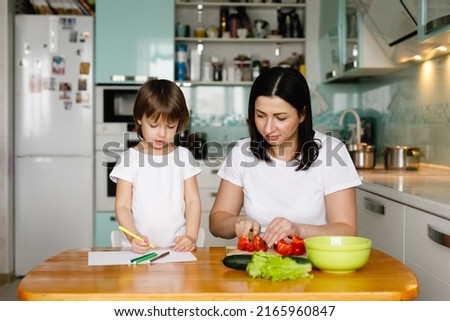 Mother and little daughter sitting in the kitchen, making vegetable salad, drawing. Happy family concept. Daily home life