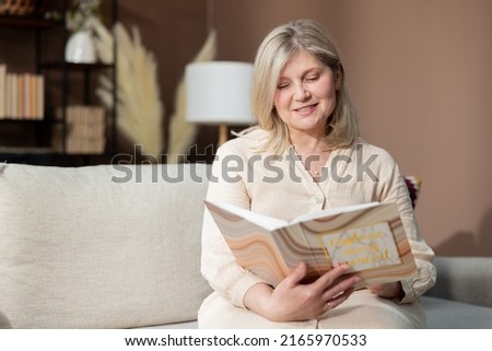 An elderly beautiful smiling woman holding a photo album sits on a comfortable sofa in the living room happily looking at family photographs of vacation memories.