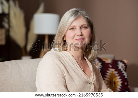 Portrait of beautiful smiling woman with blonde gray hair relaxing in living room on couch. Older mature complexion of elegant retired senior lady.
