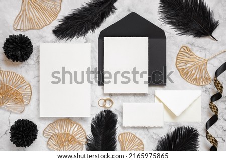 Blank cards and black envelope on marble table near black feathers and golden leaves top view. Wedding set mockup. Elegant femminine suite with paper cards, flat lay 