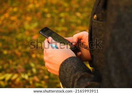 Defocus male hand holding phone. Man using smart phone in autumn park. Typing text message or reading social media at mobile phone. Fall background. Cell phone. Communication. Out of focus.