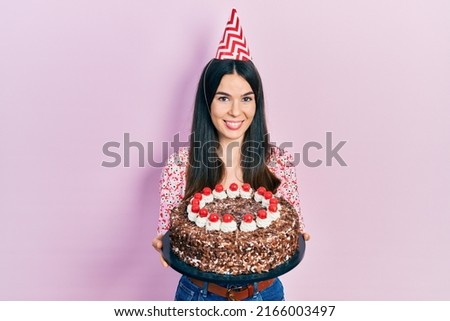 Young brunette woman celebrating birthday holding big chocolate cake smiling with a happy and cool smile on face. showing teeth. 