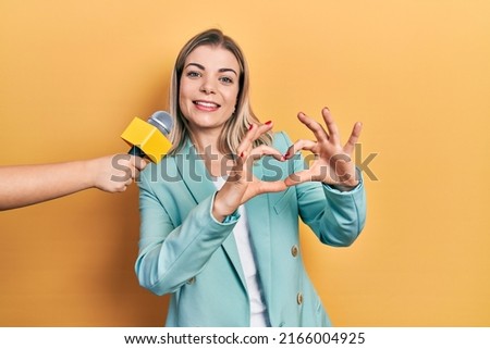 Beautiful caucasian woman being interviewed by reporter holding microphone smiling in love showing heart symbol and shape with hands. romantic concept. 