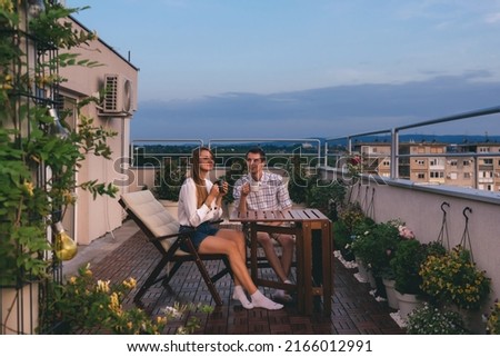 Young couple relaxing outdoors on urban rooftop garden with blooming flowers. Man and woman in casual clothes enjoying sunset and drinking tea or coffee at apartment balcony terrace with city view. Royalty-Free Stock Photo #2166012991