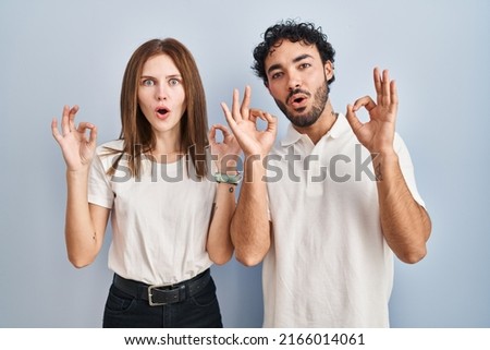 Young couple wearing casual clothes standing together looking surprised and shocked doing ok approval symbol with fingers. 