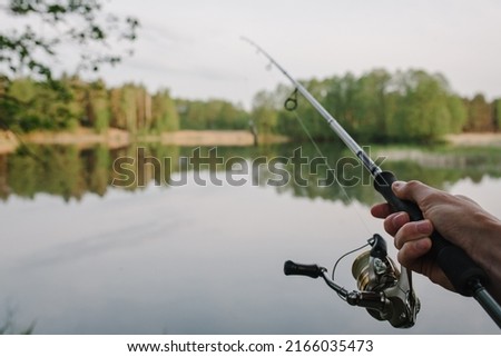 Man catching fish, pulling rod while fishing from lake or pond. Fisherman with rod, spinning reel on river bank. Sunrise. Fishing for pike, perch, carp on beach lake or pond. Background wild nature. Royalty-Free Stock Photo #2166035473