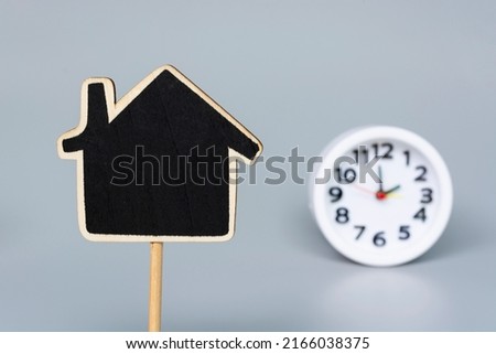Wooden house model with blurred white alarm clock background. Real estate investment concept. Copy space.