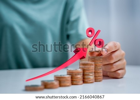 Woman's hand placing coin on stack of coins arranged in upward position and illustration of red arrow pointing upward indicating rising interest rates. Royalty-Free Stock Photo #2166040867