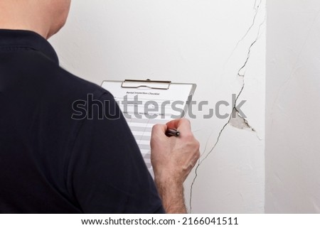 Man with inspection checklist in front of a white wall with a long crack or rip and a piece of plaster missing, rental damage concept. Royalty-Free Stock Photo #2166041511