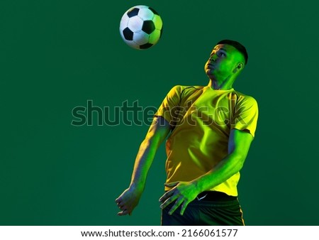Half-length of professional male football soccer player playing with ball isolated on green background in neon light. Concept of sport, goals, competition, game, achievements. Man in yellow football