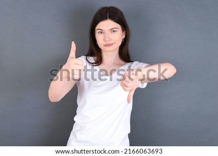 beautiful Caucasian woman wearing white T-shirt over studio grey wall feeling unsure making good bad sign. Displeased and unimpressed.