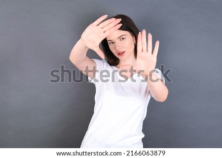 Portrait of smiling beautiful Caucasian woman wearing white T-shirt over studio grey wall looking at camera and gesturing finger frame. Creativity and photography concept.