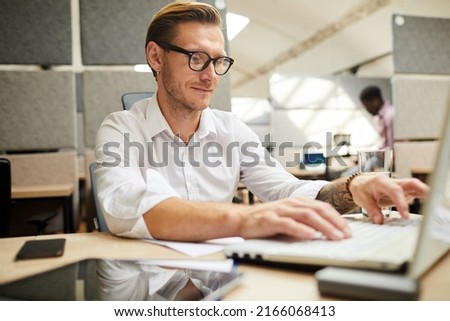 Content creative man with tattoo sitting at table in office of marketing company and editing presentation on laptop