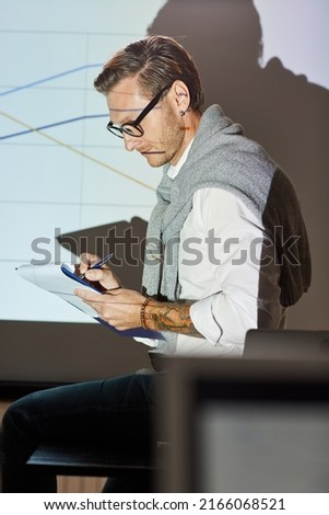 Content experienced financial coach in smart casual outfit sitting on table against projection screen and making notes in paper