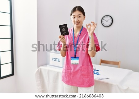 Young asian nurse woman at political campaign election holding usa passport doing ok sign with fingers, smiling friendly gesturing excellent symbol 