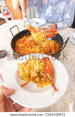 Woman's hands taking a picture of a rice paella with lobster. Older woman's hand in the background wants to take a glass of wine. A young woman hands wants to take the empty plates to serve the rice. 
