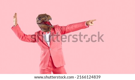 Funny goofy guy having fun in the studio. Happy energetic excited man wearing stylish funky vibrant pink party suit and dinosaur mask dancing and pointing finger away isolated on pink background