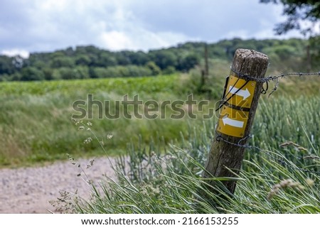 Wooden post with two yellow plaque, arrows pointing the way, hiking trail between Dutch farmland meadows, lush trees in blurred background, spring day in Beek, South Limburg, Netherlands
