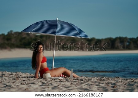Young Latina girl lying on the beach in summer looking at the camera with an umbrella. With a hat, a book and a soft drink beside her. Horizontal photo