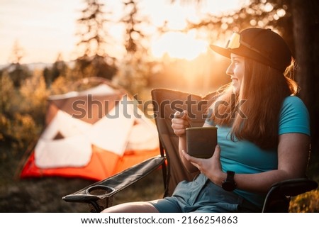Woman sitting in chairs outside the tent. Sunset camping in forest. Recreation outdoor activity. Cooking dinner with camping gear in camp. Summer travel outdoor adventure