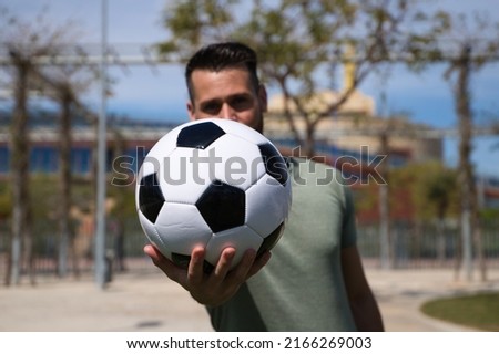 Handsome young man holds a football in his hand and shows it to the camera in focus. The man is a sports fan and in particular a football fan. He is a fan of his team. Concept of sports and team play.