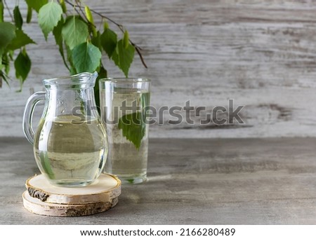Birch sap in a transparent jug on a wooden stand. A tall glass of sap and a sprig of birch in the background. Light wooden background, space for text Royalty-Free Stock Photo #2166280489