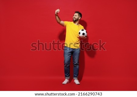 Full size body length young bearded man football fan in yellow t-shirt support favorite team hold soccer ball do selfie shot on mobile cell phone isolated on plain dark red background studio portrait