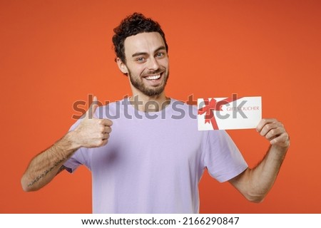 Smiling young bearded man 20s wearing casual basic violet t-shirt standing hold gift certificate showing thumb up isolated on bright orange color wall background studio portrait. Tattoo translate fun