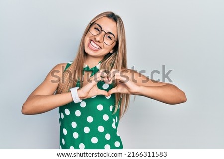 Beautiful hispanic woman wearing elegant shirt and glasses smiling in love doing heart symbol shape with hands. romantic concept. 