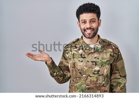 Arab man wearing camouflage army uniform smiling cheerful presenting and pointing with palm of hand looking at the camera. 