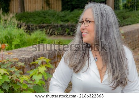 Profile portrait of a mature woman with a calm smiling expression, tanned skin, straight gray hair, glasses, light makeup, sunny day in the garden with green vegetation in the background