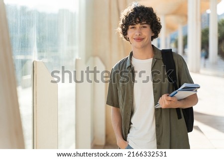 Young man is ready to travel. Happy guy with curly hair holding books and looking at the camera with while walking at the street. Stock photo