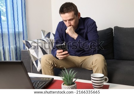 Young worried man sitting on the sofa and reading text message on mobile phone. Thoughtful serious man, looking at smartphone screen, reading bad news in message, upset male sitting on couch. Royalty-Free Stock Photo #2166335665
