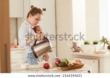 Young mother making hot tea standing at cooking table in kitchen while holding her baby Royalty-Free Stock Photo #2166344621