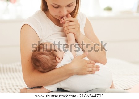 Mother smelling and kissing hand of adorable baby in bodysuit sleeping on her laps