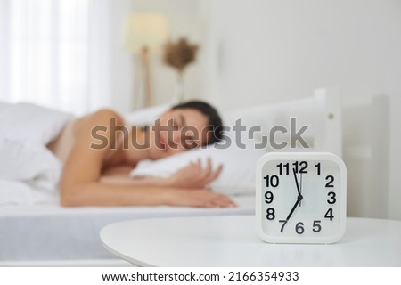 Close up of white plastic alarm clock set for 7 AM on bedside table in bedroom, with happy peaceful tranquil young woman sleeping on bed in blurred background. Sleep, morning, daily routine concepts Royalty-Free Stock Photo #2166354933