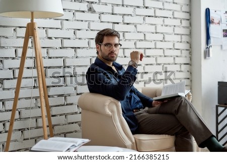 Confident caucasian IT seller look at camera while read book in armchair in office. Business strategy, development, promotion, selling and marketing of high-tech company. Successful male lifestyle