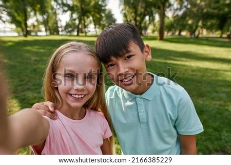 Smiling beautiful multiethnic children of generation alpha looking at camera during taking selfie on green meadow in blurred park. Boy hugging his girl friend. Childhood lifestyle. Sunny summer day