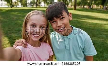 Smiling cute multiracial children of generation alpha looking at camera during taking selfie on green lawn in blurred park. Boy hugging his girl friend. Childhood lifestyle. Sunny summer day