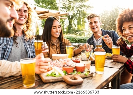 Group of multiracial friends having backyard dinner party together - Diverse young people sitting at bar table toasting beer glasses in brewery pub garden - Happy hour, lunch break and youth concept Royalty-Free Stock Photo #2166386733