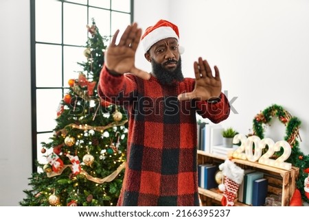 African american man wearing santa claus hat standing by christmas tree doing frame using hands palms and fingers, camera perspective 