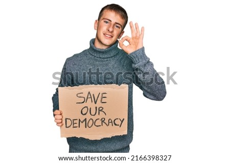 Young blond man holding save our democracy protest banner doing ok sign with fingers, smiling friendly gesturing excellent symbol 
