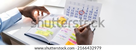Businessman's Hand Checking Schedule In Diary With Calendar On White Desk