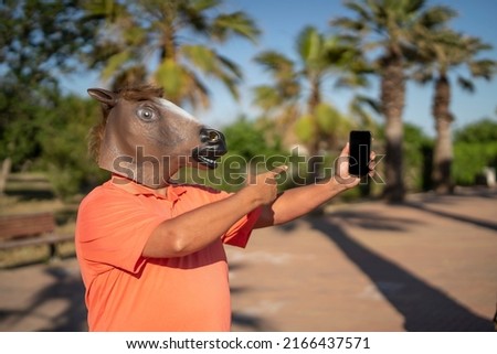 Man with horse head mask between palm trees using smartphone Royalty-Free Stock Photo #2166437571