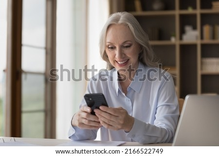Smiling middle-aged businesswoman using smartphone seated at workplace, distracted from work chatting on internet, read message from client, check calendar. Modern tech for business, agenda concept Royalty-Free Stock Photo #2166522749